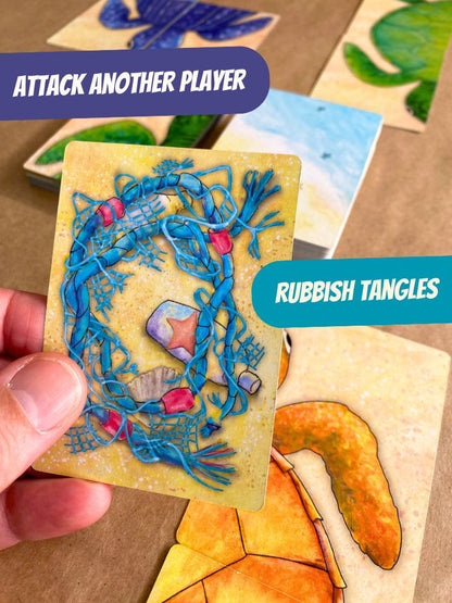 A hand holds a card titled 'Rubbish Tangles' in a card game, showing a blue tangled net illustration with small trash icons on a yellow card face. Other illustrated cards are visible in the background on a brown surface, with text overlays reading 'ATTACK ANOTHER PLAYER' and 'RUBBISH TANGLES'. In card game Turtle Moon