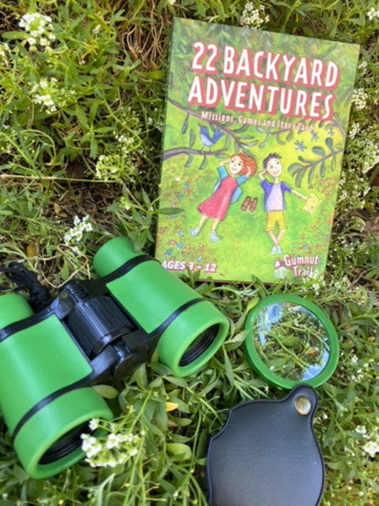 A children's outdoor-themed book titled "22 Backyard Adventures" lies on grass beside green toy binoculars, a magnifying glass, and a black accessory piece.