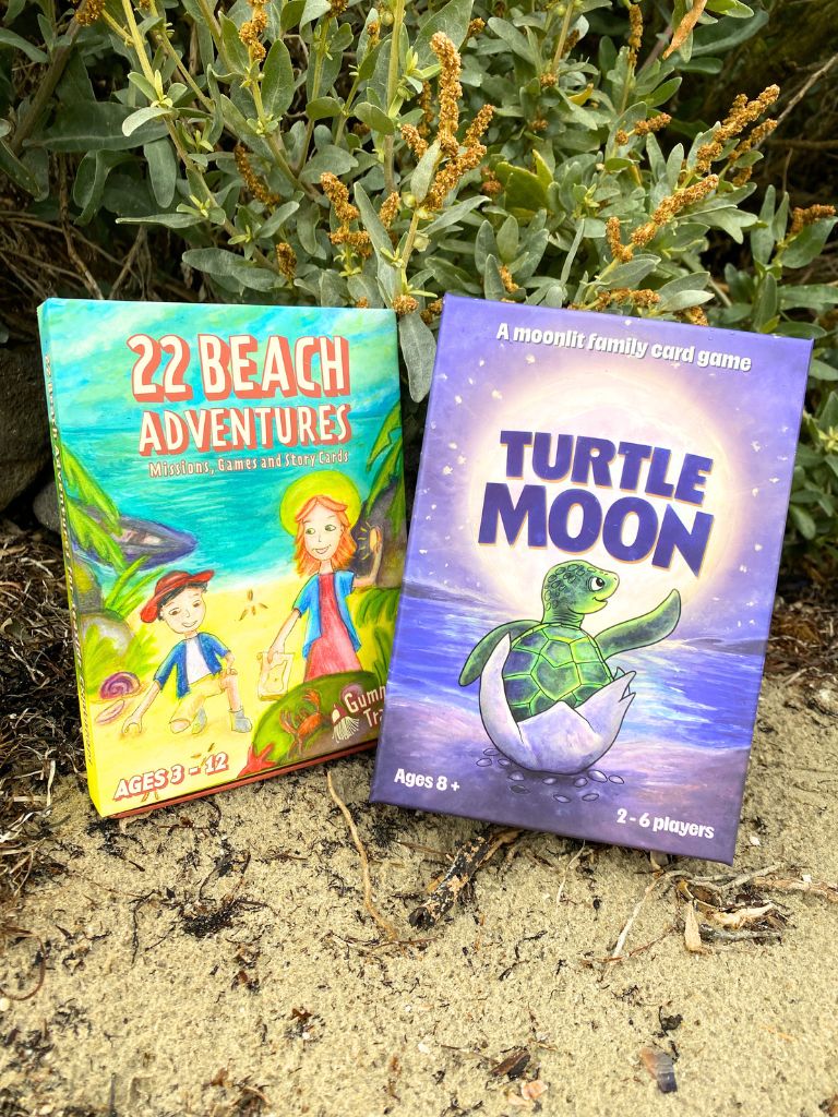 Two family friendly guides and games by Gumnut Trails standing upright outdoors on sand in front of green plants. The left box reads "22 Beach Adventures" and shows a colorful beach-themed illustration. The family card game says Turtle Moon for ages 8+