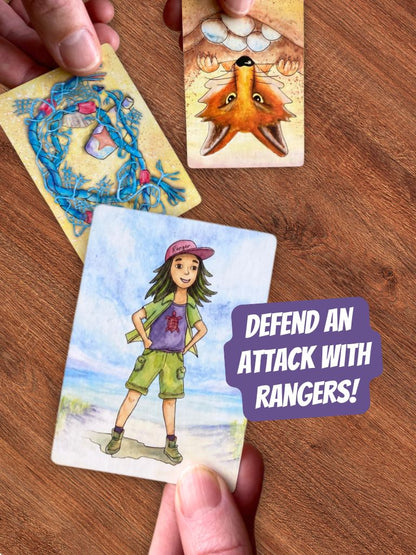 Three illustrated playing cards from Turtle Moon card game are shown on a wooden surface, with hands holding them. The front card features a girl turtle ranger wearing a pink cap and green shorts, with a purple text bubble reading 'DEFEND AN ATTACK WITH RANGERS!'.