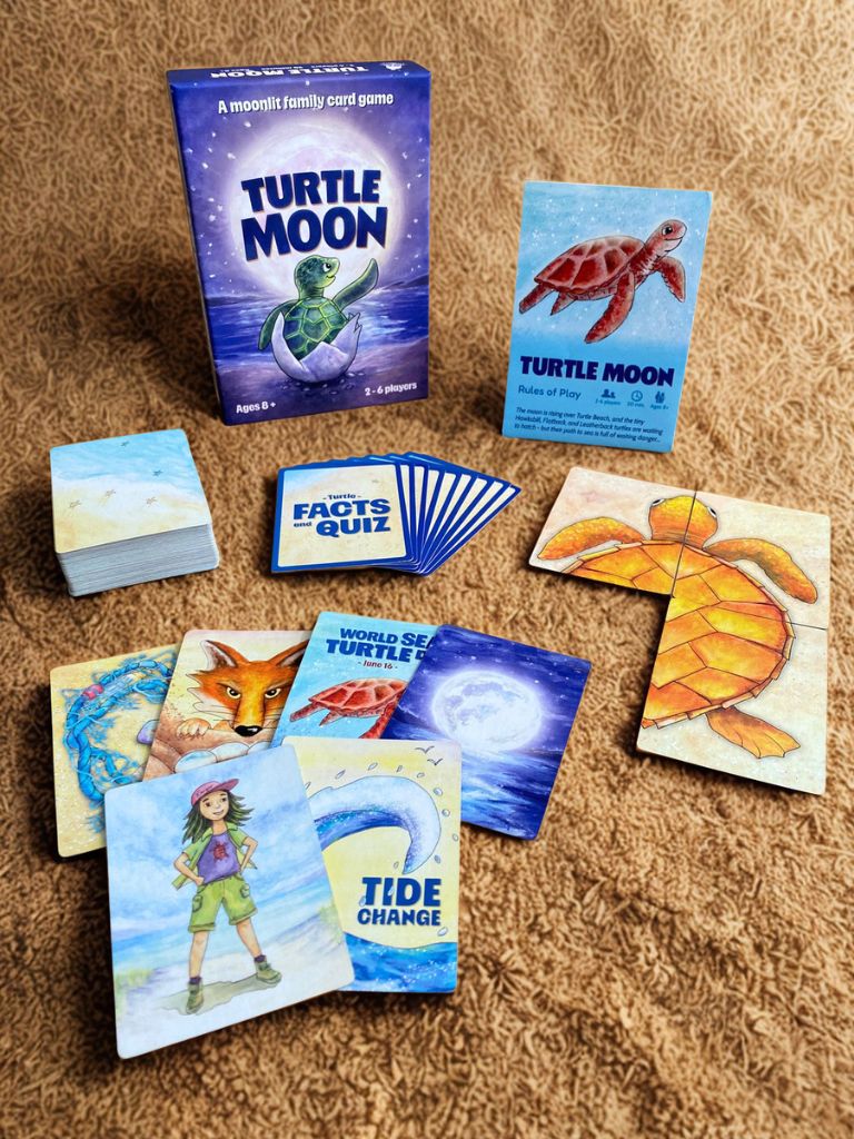 Turtle Moon family card game set arranged on a brown textured sand, including the blue game box, instruction card, facts and quiz card, and several illustrated cards showing a turtle, fox, ocean wave, and a turtle ranger. 