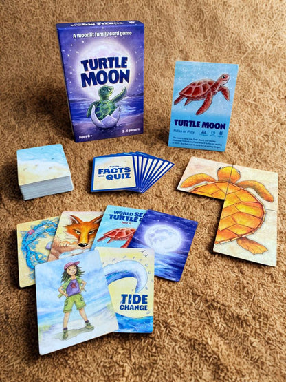 Turtle Moon family card game set arranged on a brown textured sand, including the blue game box, instruction card, facts and quiz card, and several illustrated cards showing a turtle, fox, ocean wave, and a turtle ranger. 