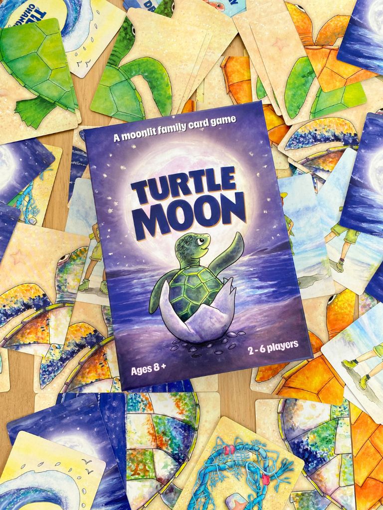 The box for the 'Turtle Moon' family card game is centered and surrounded by scattered illustrated cards on a wooden surface. The box features a purple night-sky background with a cartoon turtle hatching from an egg.