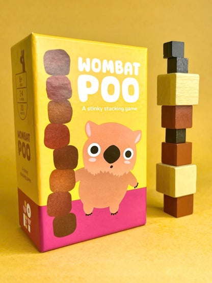 Wombat Poo board game box shown next to a stack of wooden blocks on a warm yellow background. The box is yellow and pink with a cartoon wombat illustration and the title 'WOMBAT POO' in large white letters.