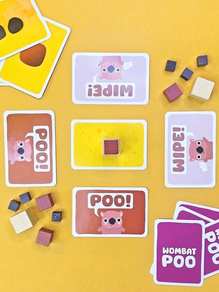 A colorful children's card game set arranged on a bright yellow background. The cards feature cartoon animal faces and text such as 'WIPE', 'POO!', 'WOMPAT POO', and 'POO!', with small wooden blocks scattered around.