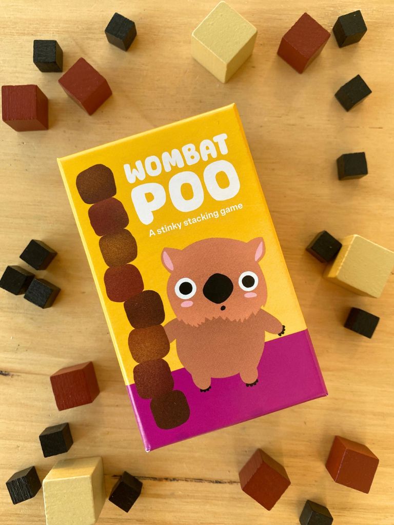 The box for the children's stacking game 'Wombat Poo' is shown on a light wooden surface, surrounded by wooden cube blocks in black, brown, and beige. The box features a bright yellow top half with the title in bold white letters, and a bottom half with a purple base and a cartoon wombat holding a stack of brown poos.