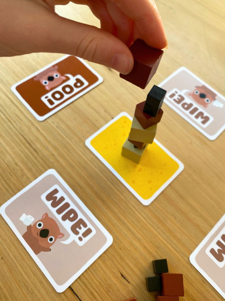 A close-up of a hand playing a wooden block toy game on a light wood tabletop. The hand is holding a dark reddish-brown block above a small tower of wooden blocks on a yellow square card, with cartoon cards reading 'POO!' and 'Wipe!' scattered around. Wombat Poo Game