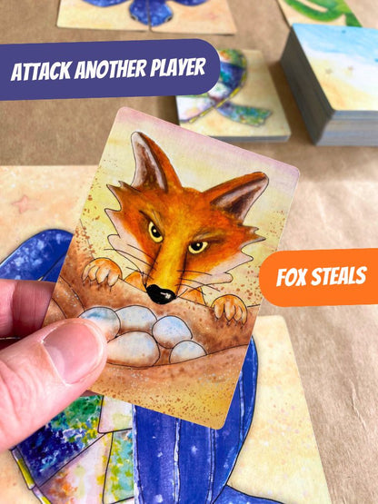A hand holds a fox card in Turtle Moon with yellow eyes peering over blue eggs, with game text overlays reading 'ATTACK ANOTHER PLAYER' and 'FOX STEALS'.