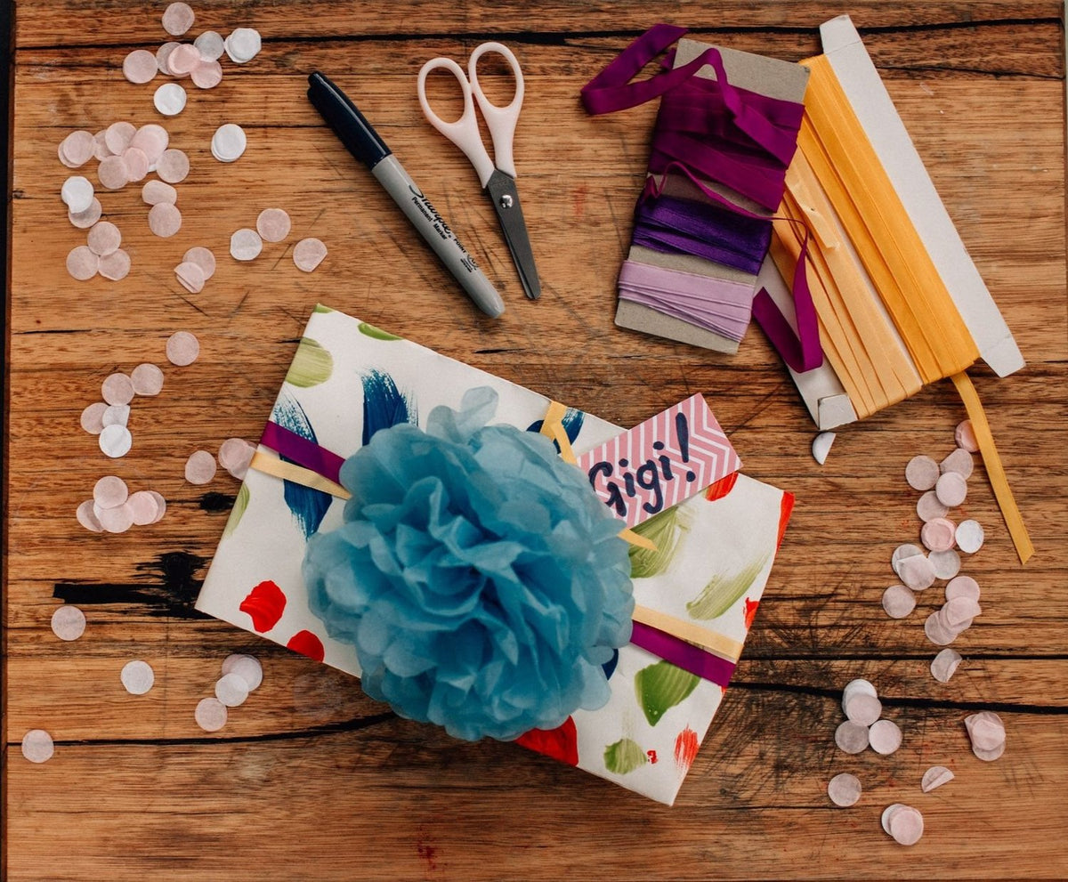 A wrapped gift box on a rustic wooden table, decorated with a large blue tissue flower, a colorful patterned paper wrap, a pink chevron tag, and surrounded by craft supplies including scissors, markers, ribbon, and paper fans.