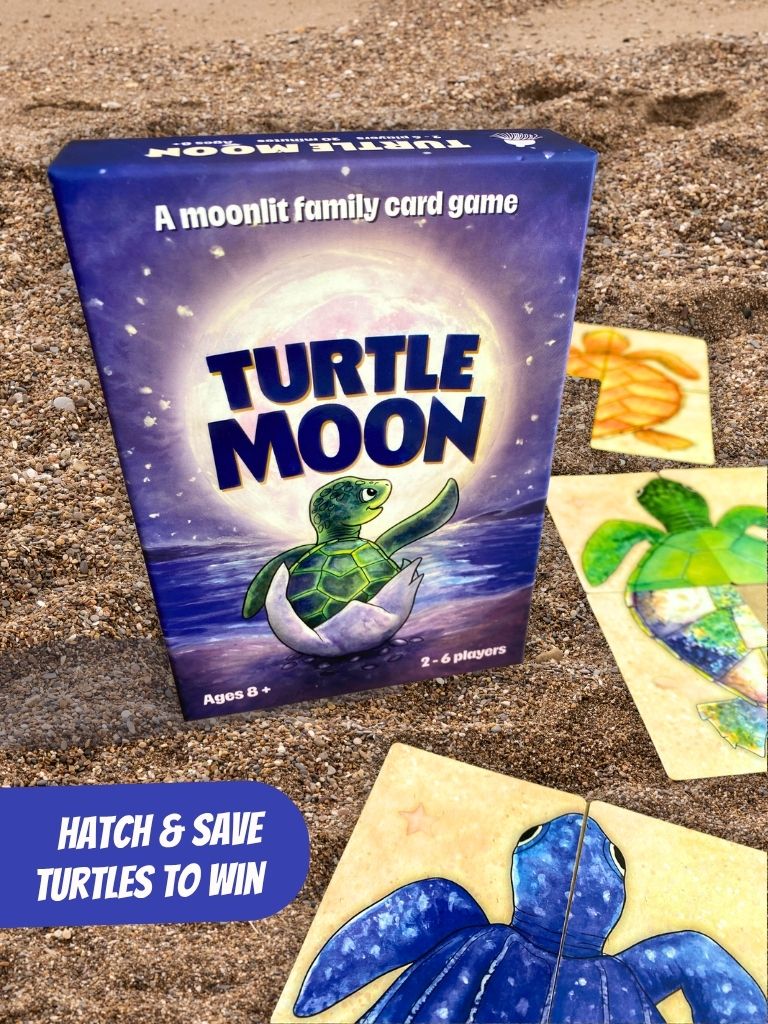 Turtle Moon family card game box displayed on a sandy surface with several turtle-themed cards scattered around it. The box features a blue and purple night-sky design with a green turtle hatching from an egg, and the text 'A moonlit family card game' and 'TURTLE MOON'.
