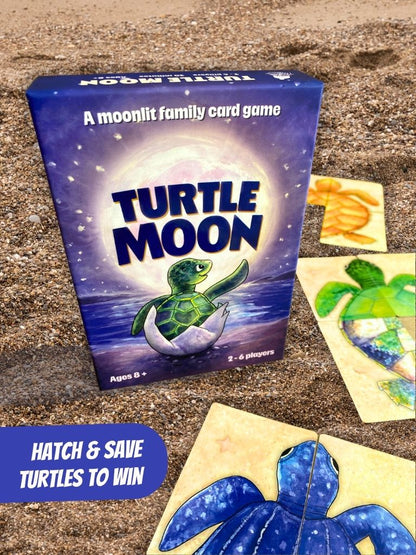 Turtle Moon family card game box displayed on a sandy surface with several turtle-themed cards scattered around it. The box features a blue and purple night-sky design with a green turtle hatching from an egg, and the text 'A moonlit family card game' and 'TURTLE MOON'.