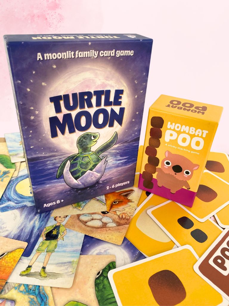 Turtle Moon family card game set with the blue game box, a yellow Wombat Poo card deck box, and several illustrated cards spread out on a pale pink background.