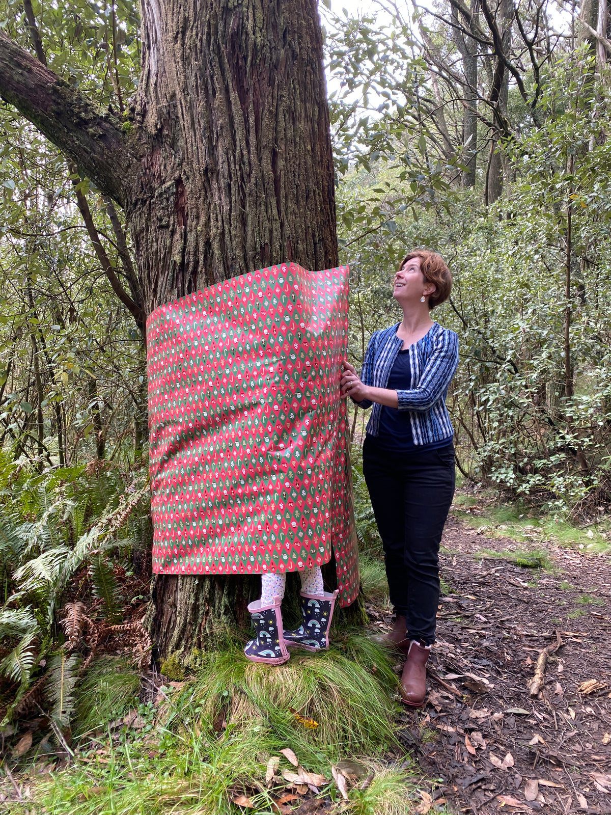 A woman stands beside a large tree in a forest, holding up a red and green patterned fabric panel that partially covers the tree trunk. The panel has a repeating floral or leaf-like print in red, green, and white.
